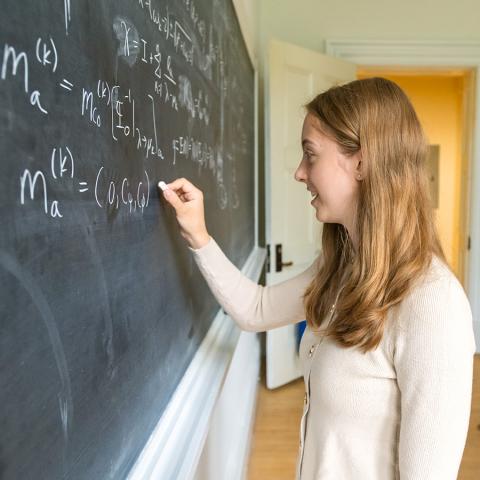 A person writing a long mathematical equation with chalk on a blackboard.