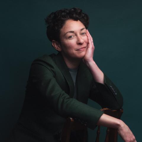 A person with short, dark curly hair wearing a dark green jacket rests their cheek against one hand while leaning over the back of a wooden chair against a solid dark green backdrop.