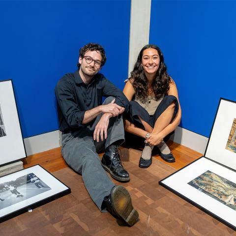 Two people sit on the floor of a gallery next to framed artworks that will soon be hung on the walls.