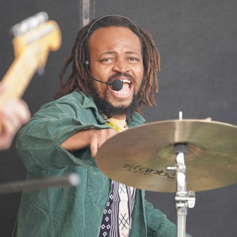 A person with dreadlocks and a headset microphone has their mouth open while behind a drum set cymbal, wearing a green corduroy jacket over a patterned top. A guitar headstock is partially visible on the left.