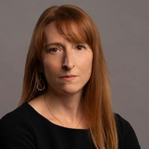 A person with shoulder-length reddish-brown hair and bangs looks directly forward against a solid gray backdrop, wearing a black top, a thin gold necklace, and a gold teardrop earring.