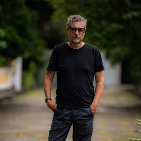 A person with short gray hair, facial hair, and black-framed glasses stands outdoors with hands in pockets, wearing a black t-shirt and dark pants against a blurred background of greenery.