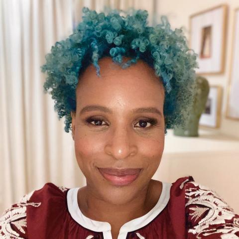 A person with short, bright blue curly hair smiles at the camera while wearing a maroon top with white embroidery. Light curtains and framed wall art are visible in the background.