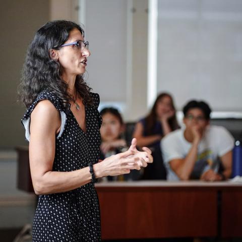 A person wearing glasses and a sleeveless dress speaking to a group of students.