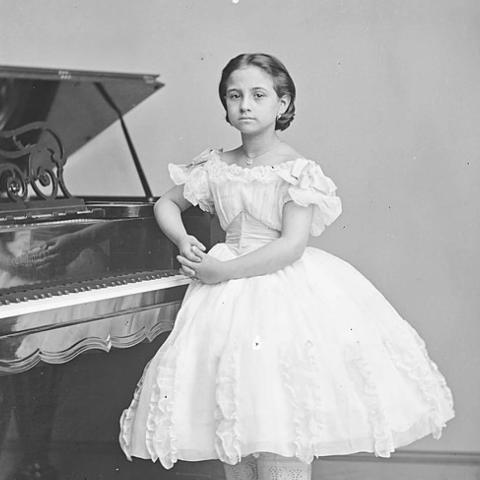 A person stands beside an open grand piano, wearing a full-skirted, off-the-shoulder white dress with lace-trimmed pantalettes and dark boots. The individual poses with hands clasped in front against a plain wall and patterned rug.