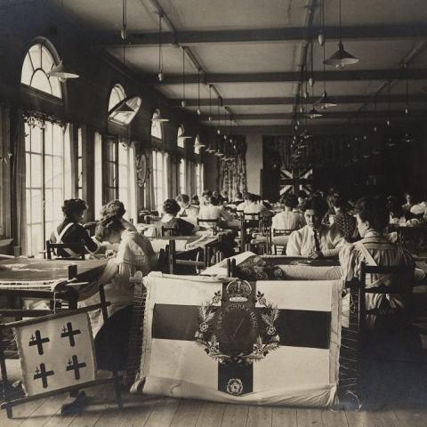 Early 20th century photograph of a large room where dozens of women sit working around needlework looms. Two flag-like needlework pictures are displayed in the foreground. 