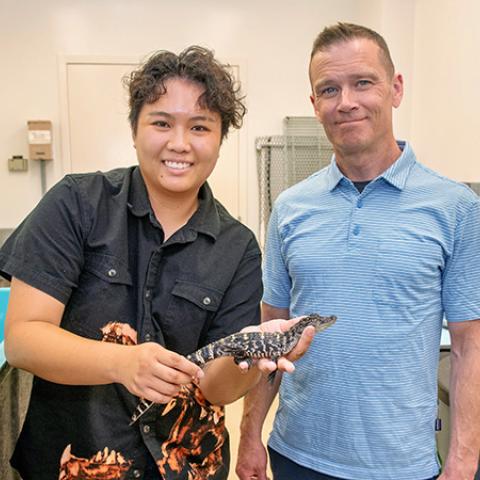 Two researchers stand in a laboratory, one holding a baby alligator beside a large turquoise tank.