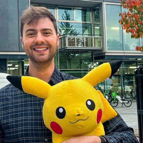 Person smiling while standing in front of a building and holding a Pokémon character.