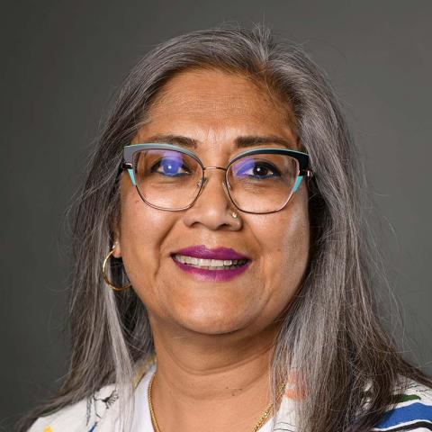 A smiling person with long gray hair and glasses against a dark gray background.
