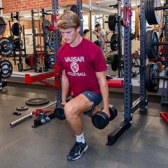 Male student wearing red shirt and gray shorts using hand weights in front of training equipment