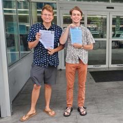 Christopher Unruh ’23 wearing navy blue patterned shirt, gray shorts and tan sandals and Chase Engel ’23 wearing gray and white patterned shirt, orange pants and brown sandals holding a document with immigrants’ legal rights in front of the doors of a  Goodwill store.