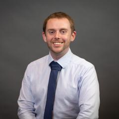 Person with short brown hair in a white collared shirt and blue tie smiling for a portrait,