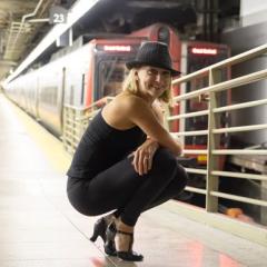 A photo of Lisa Harvie, a person with blond hair and a black fedora, crouching on a train platform.