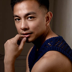 A portrait of Norbert de la Cruz III. They are seated in a three-quarter pose, turned slightly to the side with one hand resting against their chin. They have short dark hair and wear a sleeveless, dark blue knitted top. The lighting is soft and directional, casting gentle shadows that define the contours of the face and shoulder. The background is plain and dimly lit, keeping the focus on the subject.