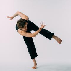 A person in a sleeveless black outfit performs a dynamic dance pose against a seamless white background. They balance on one foot with the other leg lifted and bent, while their torso leans forward and one arm extends outward in a curved motion. The fingers on one hand are splayed in a deliberate gesture, adding to the sense of motion. The lighting is even and soft, highlighting muscle definition and body alignment.
