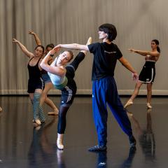 Dancers rehearsing in a studio, with one dancer extending in a supported pose while others practice movements in the background.
