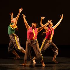 Three spotlit dancers strike poses with arms raised during a performance on a darkened stage.