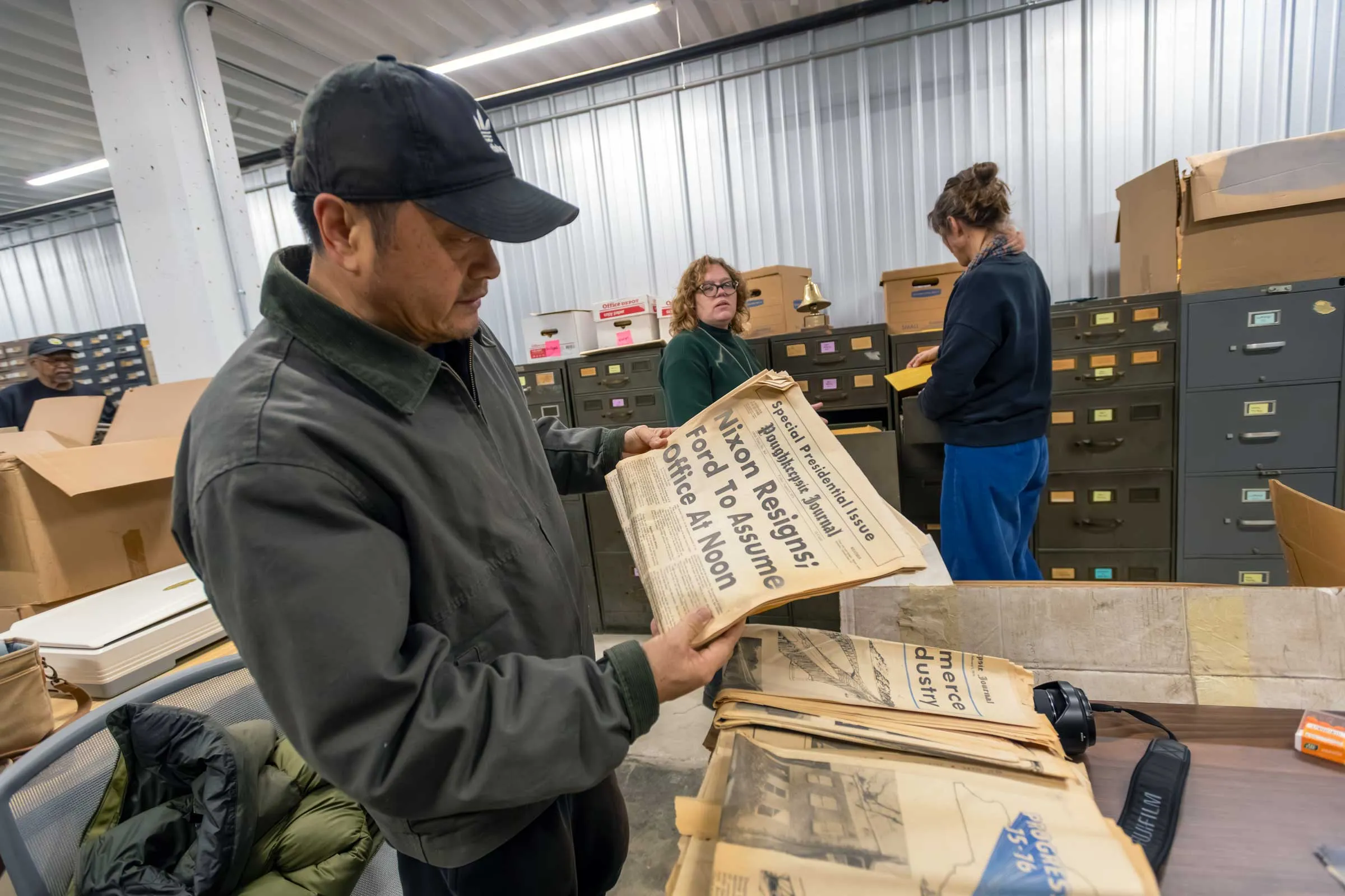 Person standing in an archive room holding and old yellowed newspaper with the front page showing.