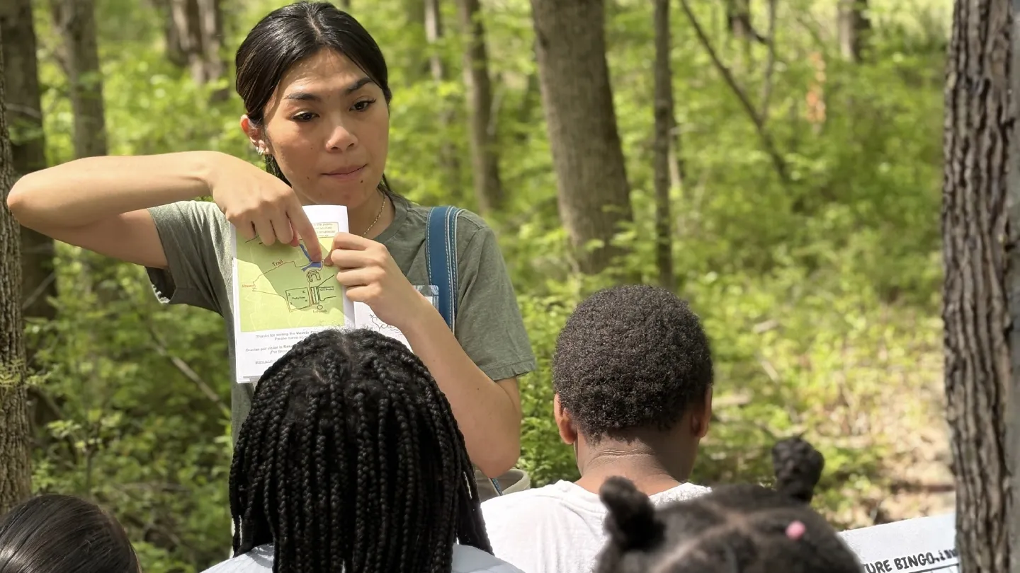 An individual stands in a wooded area, holding up a map and pointing to a specific trail location while addressing a group. The person wears a green shirt and a shoulder bag. The group, seen from behind, listens in a forest setting with green foliage.