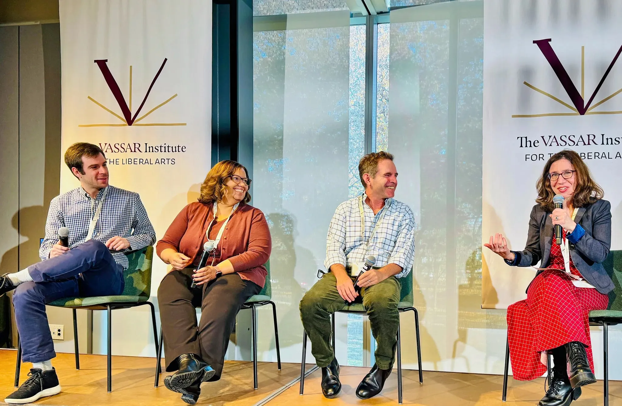 Four individuals sit on chairs arranged in a semi-circle, engaged in discussion. A backdrop featuring the Vassar Institute logo creates a formal setting, with light streaming in through large windows behind them, illuminating the scene. Each person holds a microphone; two smile while interacting, while the others appear to listen attentively. The floor is wooden, and the overall atmosphere suggests a professional yet conversational event.