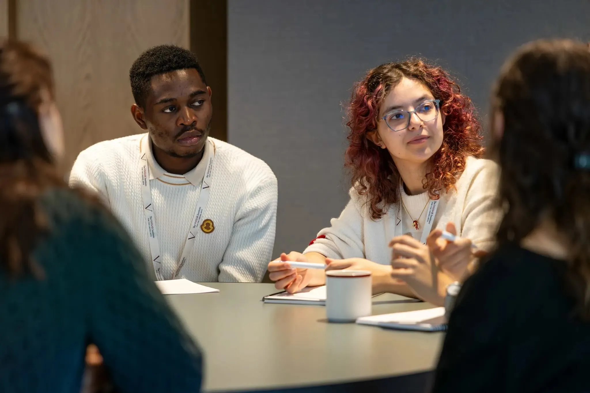 A group of four individuals is seated around a round table in a modern indoor setting. The central figures are engaged in a discussion, with one person wearing a white sweater and glasses, attentively listening, while another in a cream-colored sweater gestures as they speak. On the table, there are notebooks, pens, and a white mug, adding to the collaborative atmosphere.