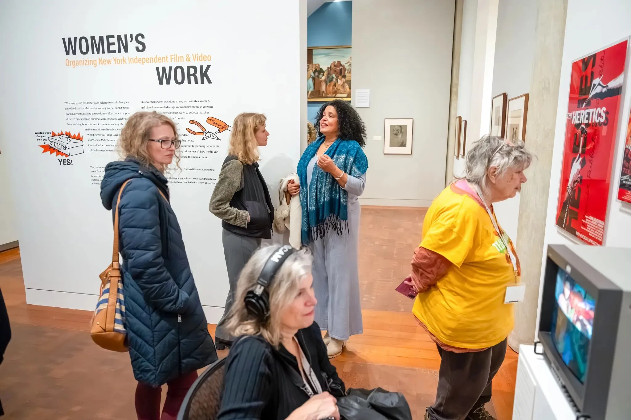 In a gallery setting, several figures are engaged with an exhibit titled "Women's Work." The wall display features text and images related to independent film and video. One individual in a blue shawl interacts animatedly, while another stands nearby, wearing a green top. To the left, a figure with headphones listens intently, and another in a yellow shirt stands near a framed poster titled "The Heretics," alongside a vintage television showing video content.