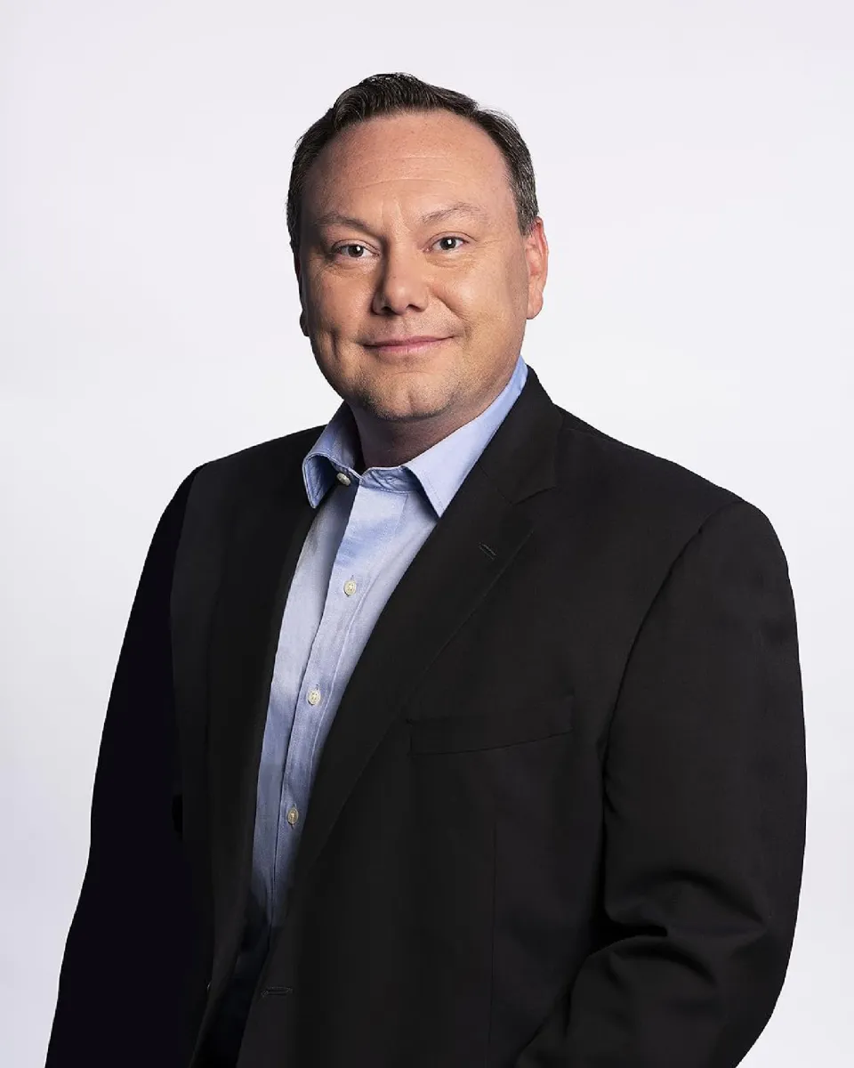 A person with short dark hair wearing a dark suit jacket and blue button-down shirt smiles at the viewer.