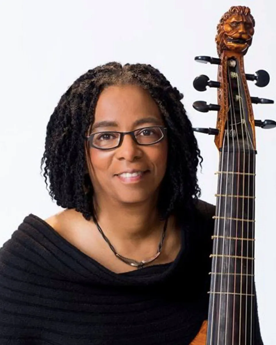 Patricia Ann Neely ’75, a person with shoulder-length, coiled hair and glasses wearing a textured black garment and a necklace, sitting next to a tall, stringed musical instrument with an intricately carved scroll shaped like a lion’s head.