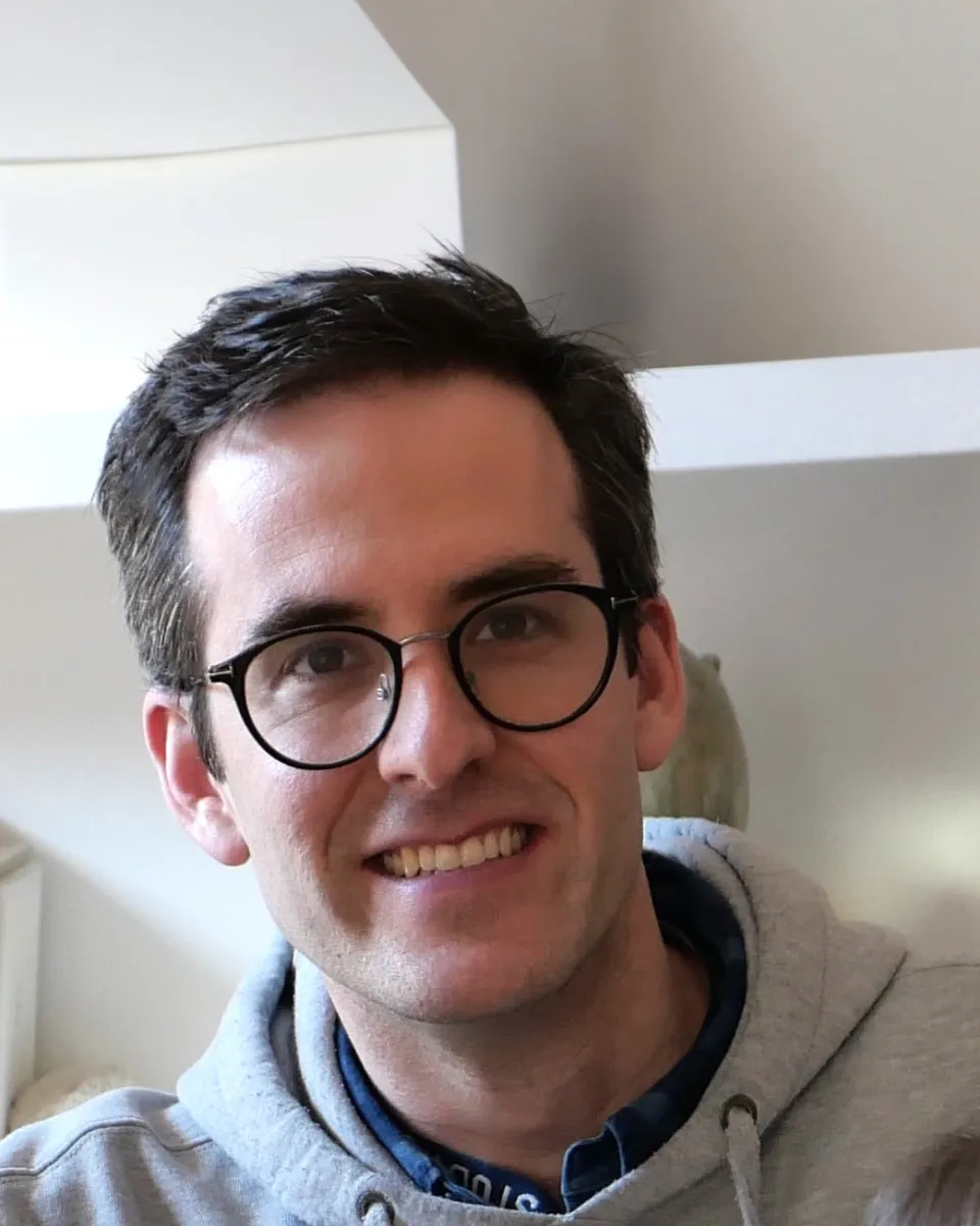Sam Thypin-Bermeo ʼ11, a person with short dark hair and glasses smiling and wearing a gray hoodie layered over a dark shirt.