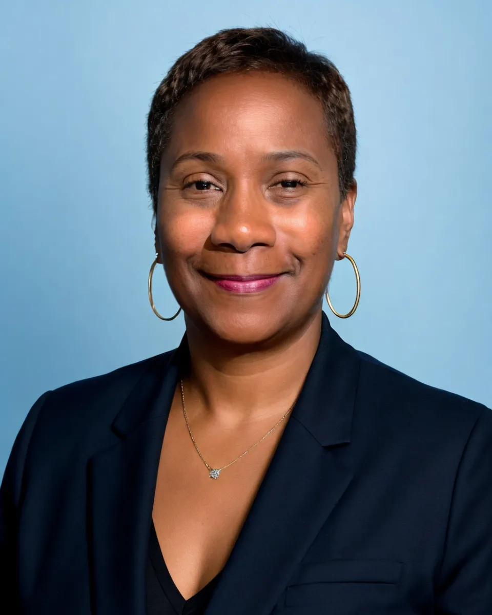 Professional headshot of a woman with short dark hair smiling at the camera. She is wearing a navy blazer over a black top, gold hoop earrings, and a delicate necklace. The background is a soft blue studio backdrop.